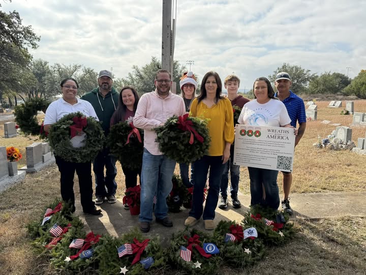 Volunteers at the flagpole holding balsam wreaths and a Wreaths Across America sign, with the service wreaths laid out on the ground before them.